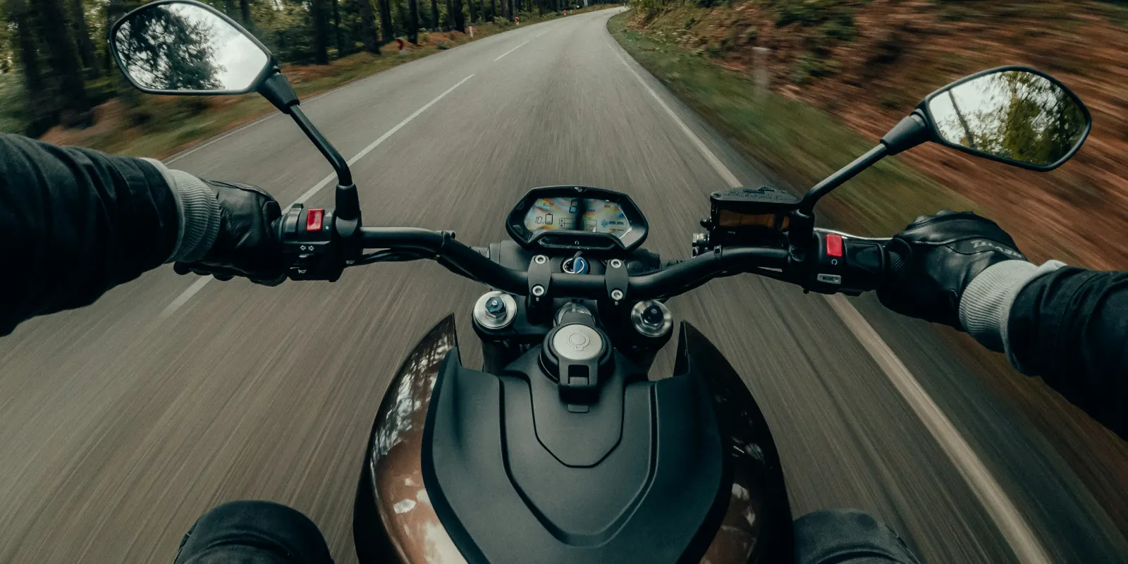Vue du pilote sur le guidon d’une moto, levier d’embrayage et commodos, sur une route en forêt